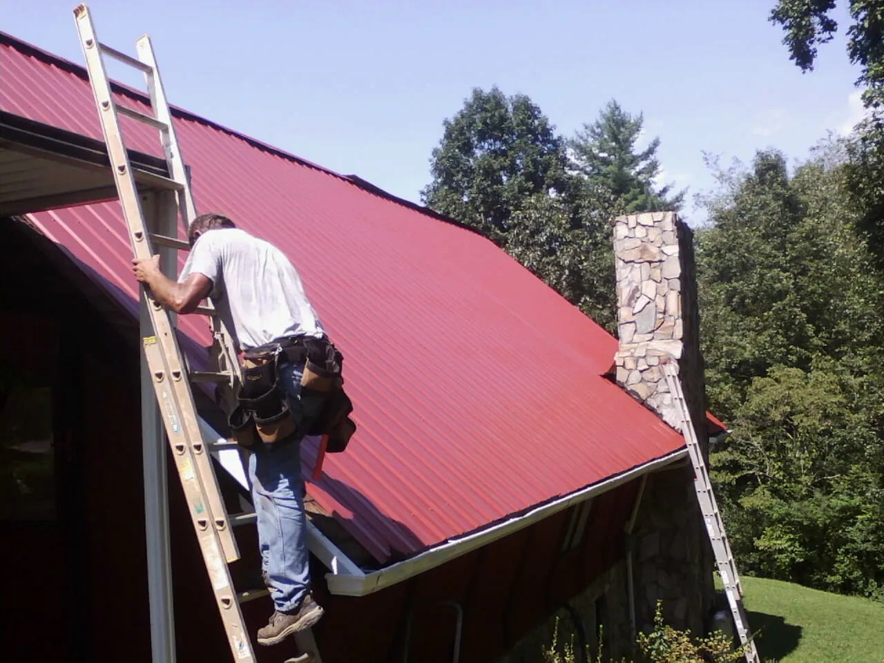 Roofer on a ladder beside a red metal roof and stone chimney — Soffit & Fascia Repair by Roofing Brothers in Coburn