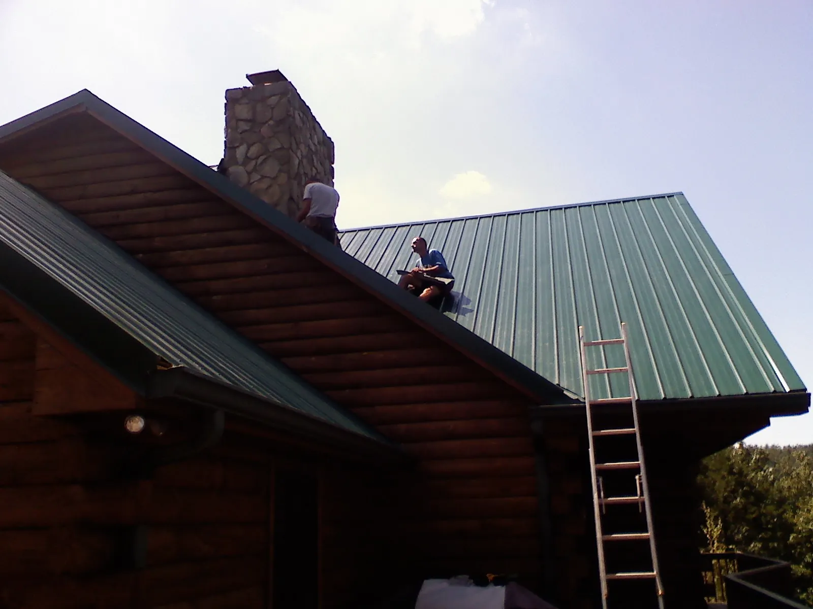 Roofers installing green metal panels beside a stone chimney on a log home in Coburn